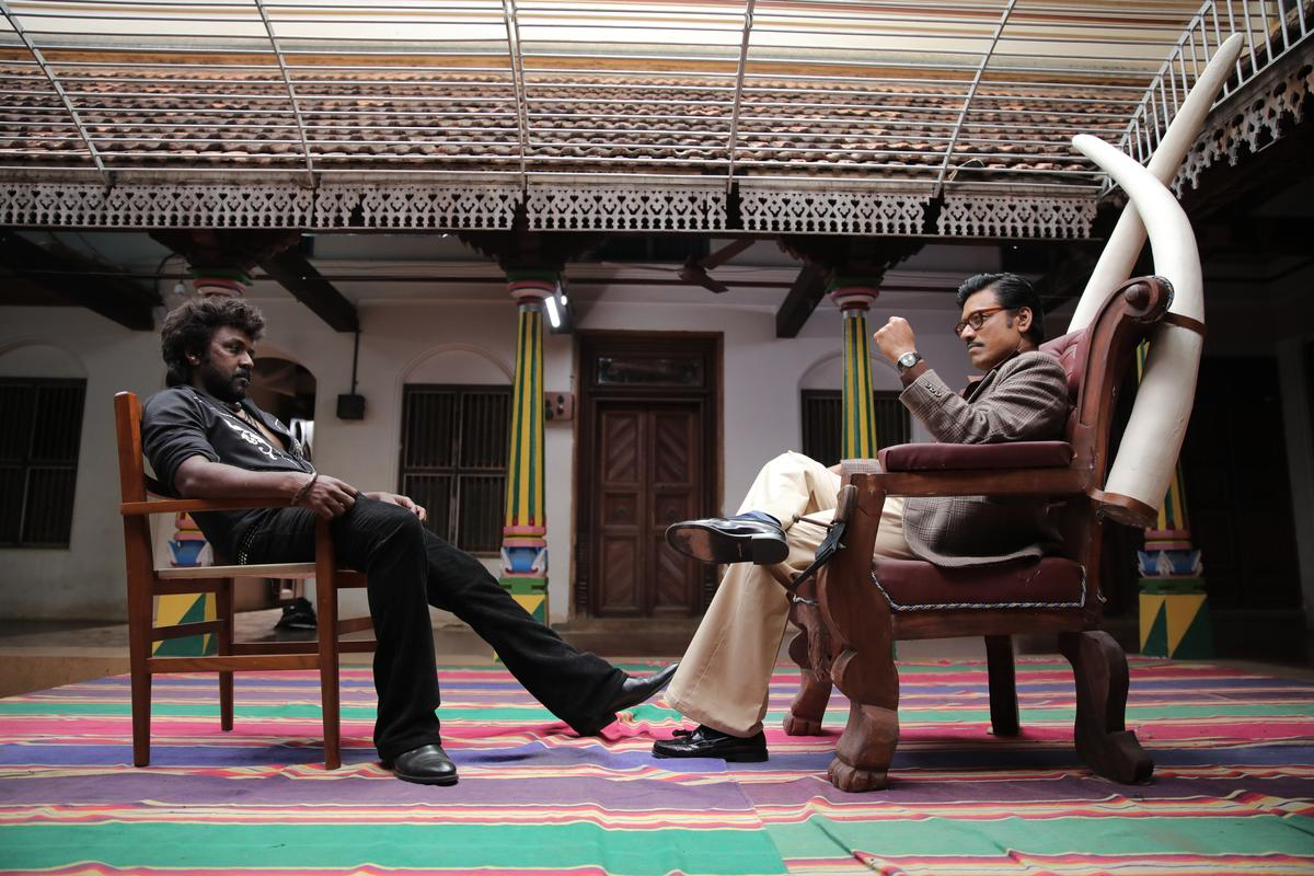 In Jigarthanda DoubleX, S.J. Suryah and Raghava Lawrence sit across from each other in chairs. Suryah's is much bigger and has giant tusks behind it.