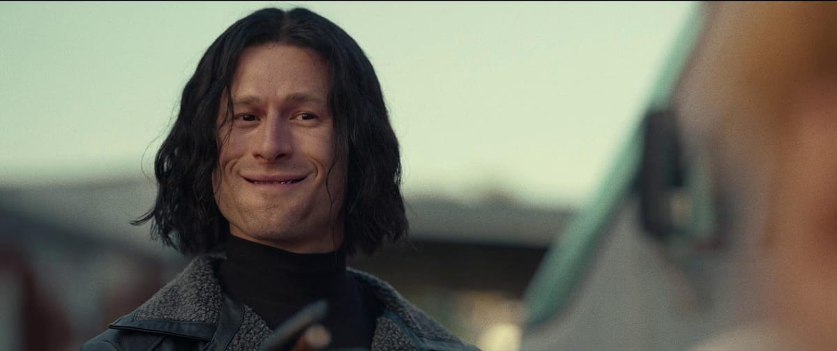Glen Powell, wearing a dark wig, gives an awkward toothy smile in Hit Man